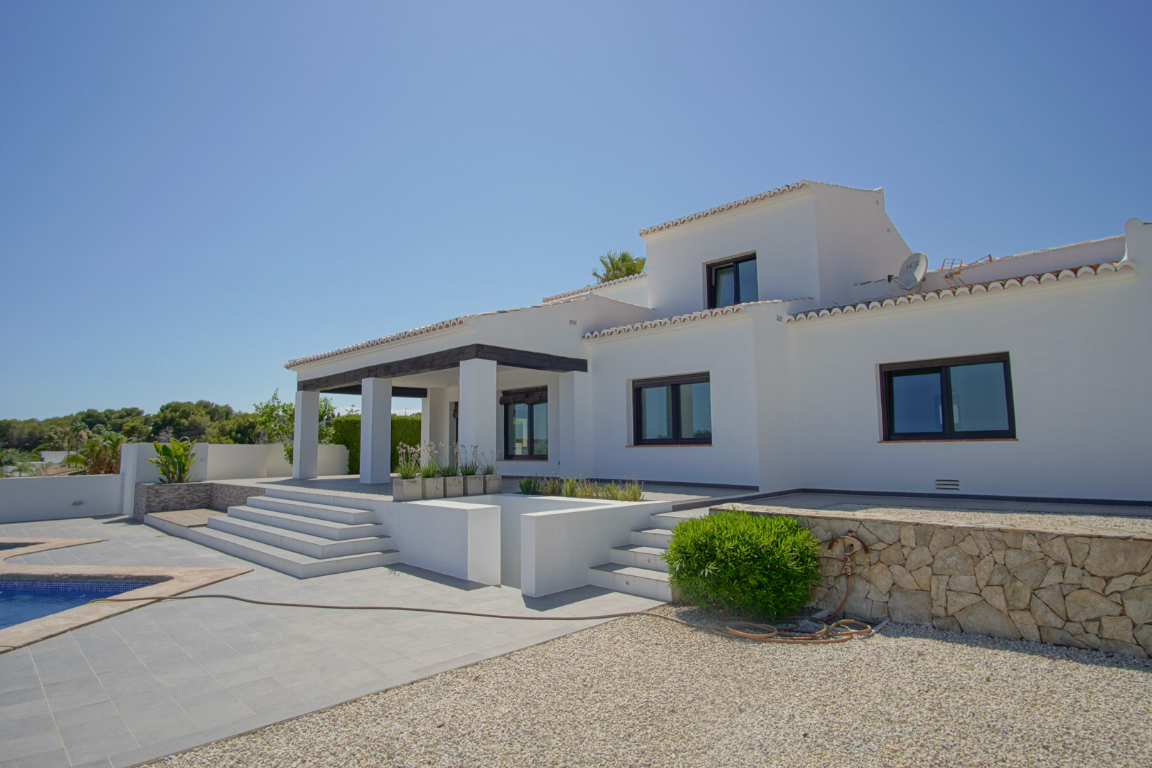 Exterior view of the Mediterranean villa featuring a white facade, dark-beamed porch, and tiered terrace connecting to the pool area and stone walls.