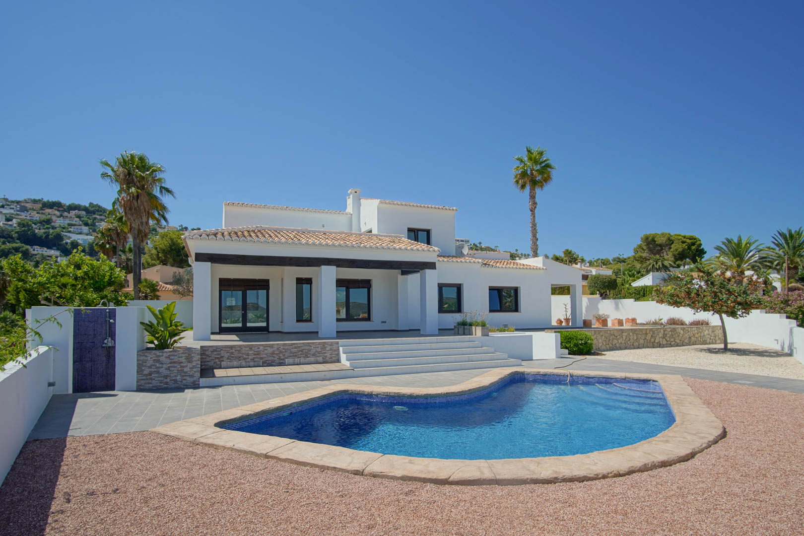 Exterior view of the villa featuring a unique-shaped pool, low-maintenance gravel gardens, and a shaded terrace with dark wooden beams and white pillars.