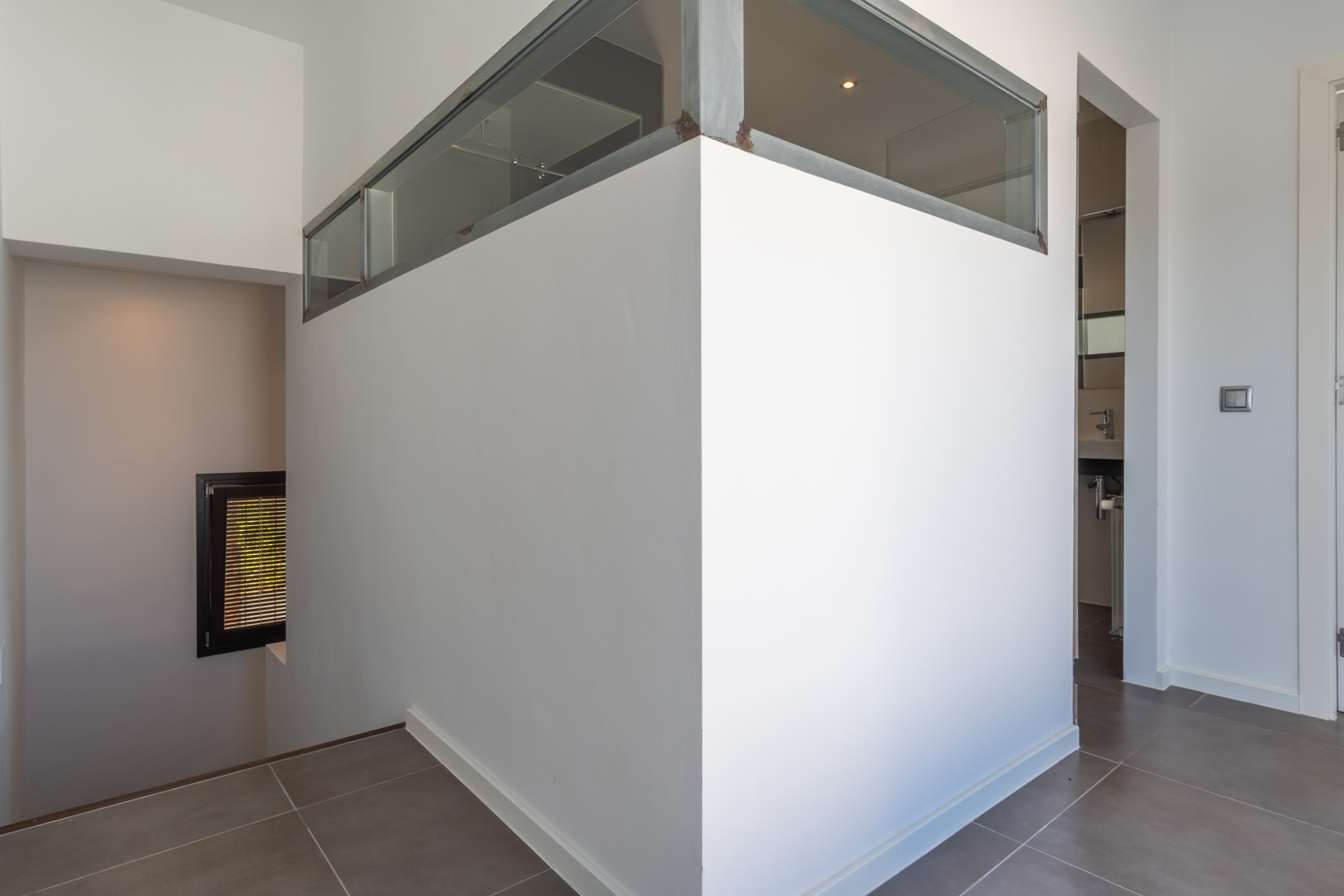 Interior hallway featuring grey floor tiles, a partition wall with industrial-style glass panels, and access to the lower level and a bathroom.