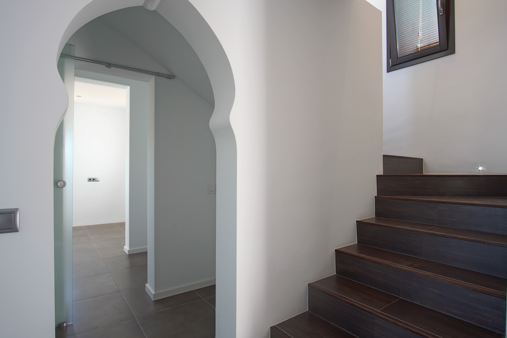 Interior staircase featuring dark tiled steps and recessed lighting, adjacent to a decorative Moorish-style archway leading to a room with a sliding glass door.