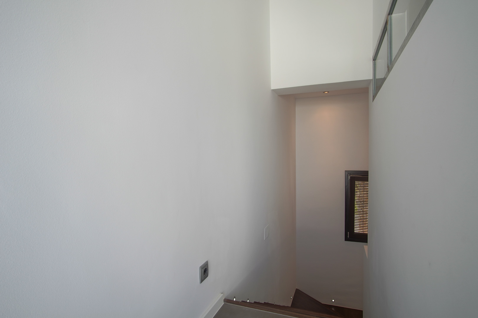 Interior staircase featuring dark wood steps and integrated floor-level LED lighting. Minimalist white walls and a small window with dark framing and shutters.