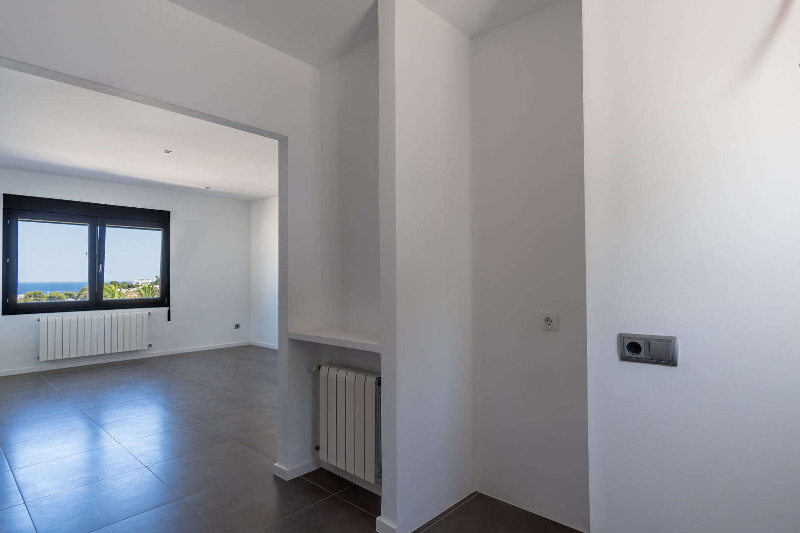 Interior view from the hallway showing grey ceramic flooring, wall radiators, and a black-framed window with views of the Mediterranean coast.