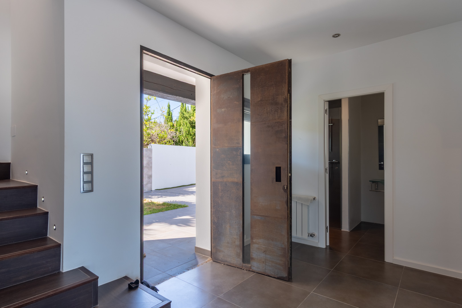 Main entrance featuring a pivot door with a corten steel finish and vertical glazing. The hall includes dark tiling, stairs with integrated LED lighting, and a bathroom.