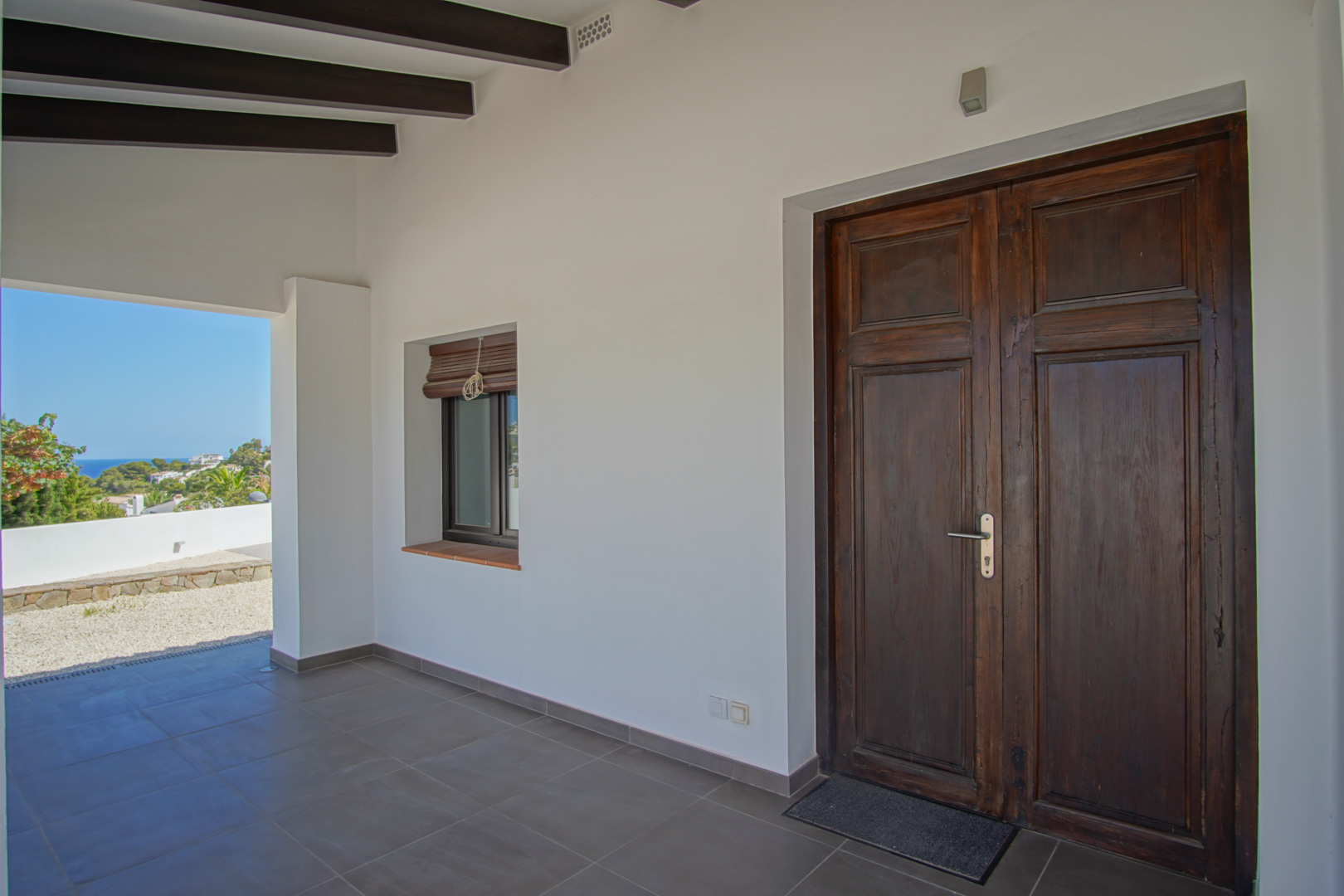 Main entrance featuring a restored antique wooden double door, dark ceiling beams, and a covered porch overlooking the Mediterranean landscape.