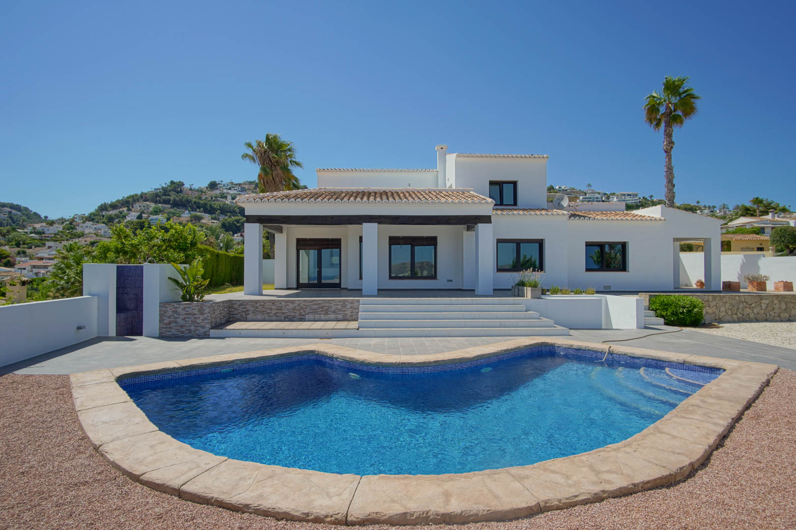 Main facade of the white Mediterranean villa featuring a curved swimming pool, covered porch with dark wooden beams, and expansive sun terraces.
