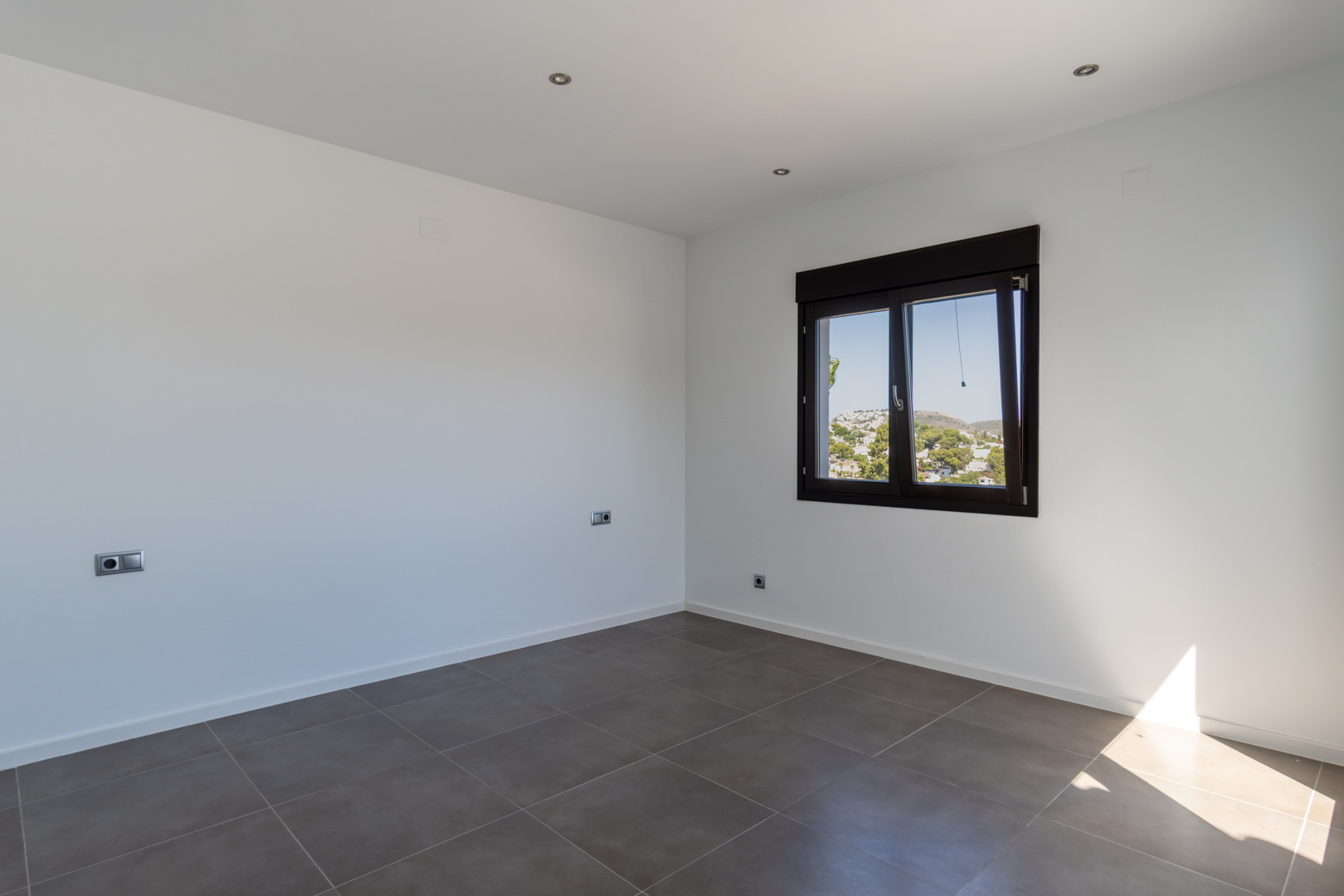 Minimalist bedroom featuring grey porcelain tile flooring, recessed LED lighting, and a dark aluminum window with views of the landscape.