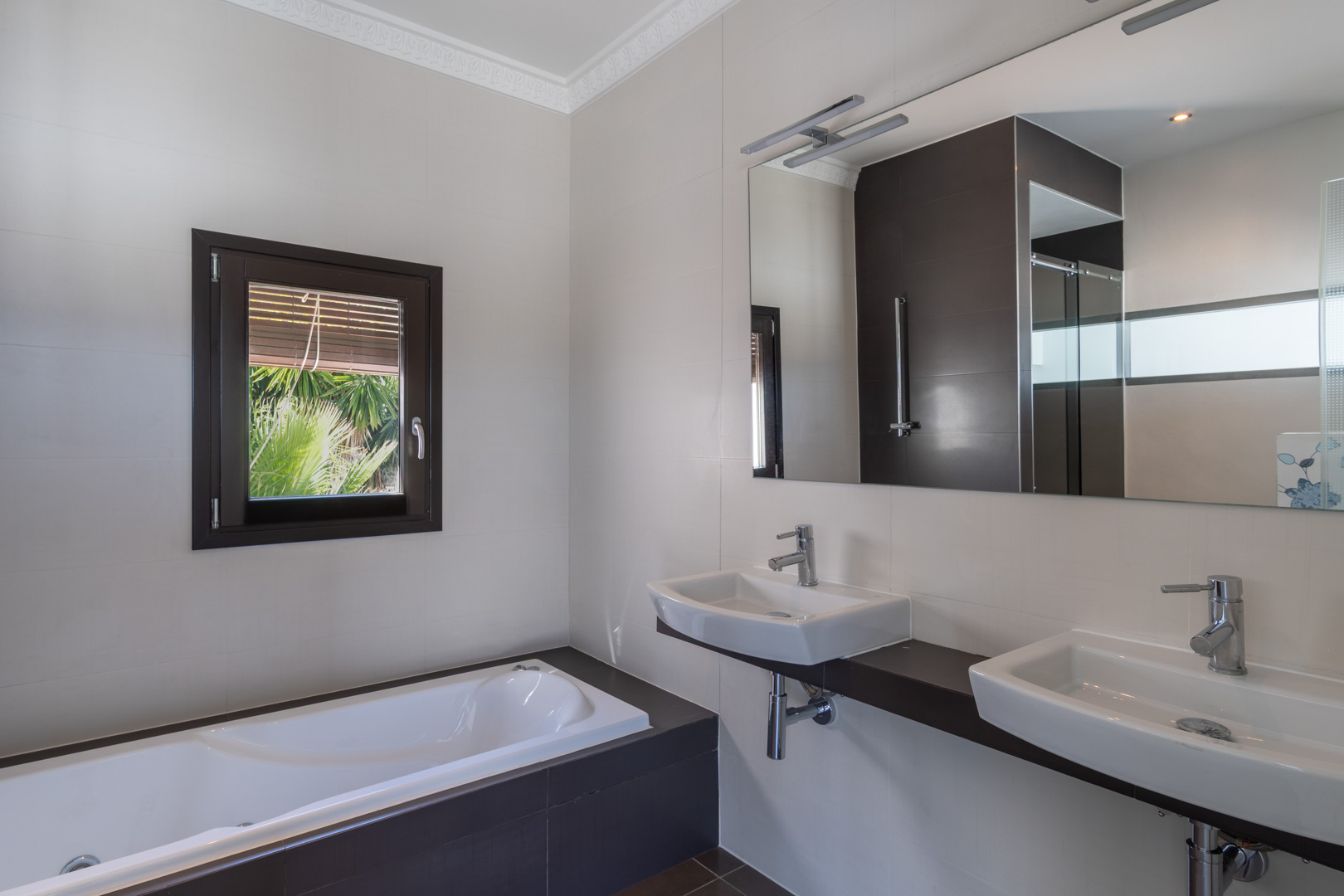 Modern bathroom featuring a double vanity on a dark shelf, an integrated bathtub, and a wood-effect window frame providing natural light and garden views.