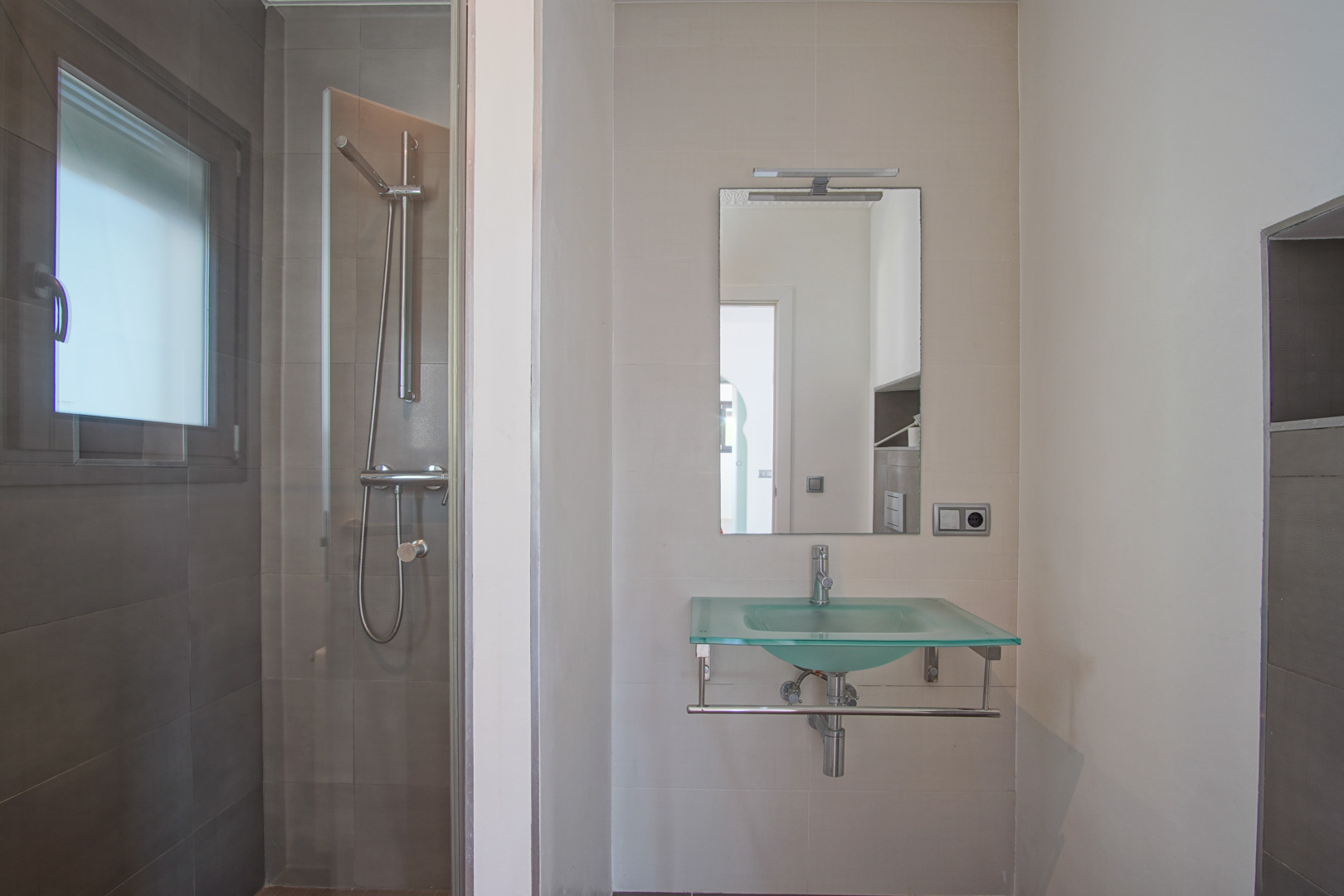 Modern bathroom featuring a translucent glass washbasin, a mirror with integrated lighting, and a walk-in shower with dark grey tiling and a glass partition.