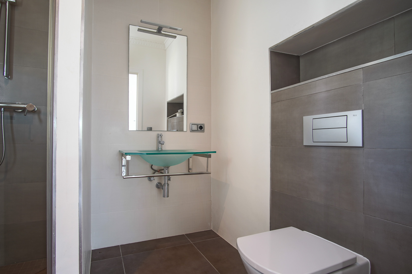 Modern bathroom featuring a wall-hung toilet, a frosted glass washbasin on chrome supports, and textured tiling with a built-in wall niche.