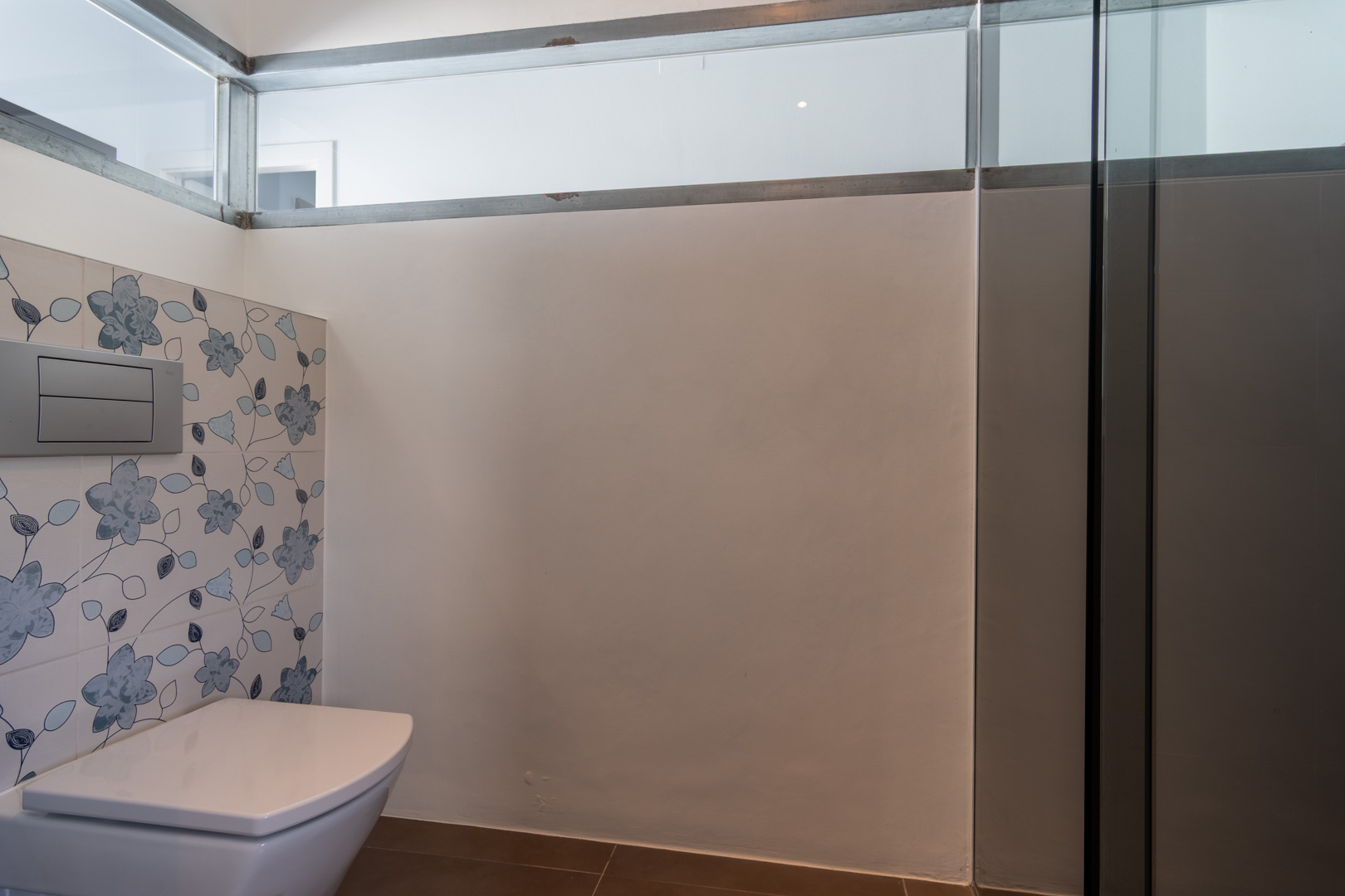Modern bathroom featuring a wall-hung toilet, decorative floral-patterned tiles in blue tones, and a clerestory glass panel for natural light.