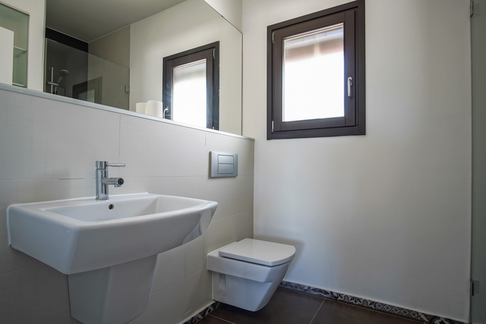 Modern bathroom featuring a wall-hung toilet, white ceramic sink, and aluminum window. It includes a large mirror and dark floor tiles with decorative hydraulic-style borders.