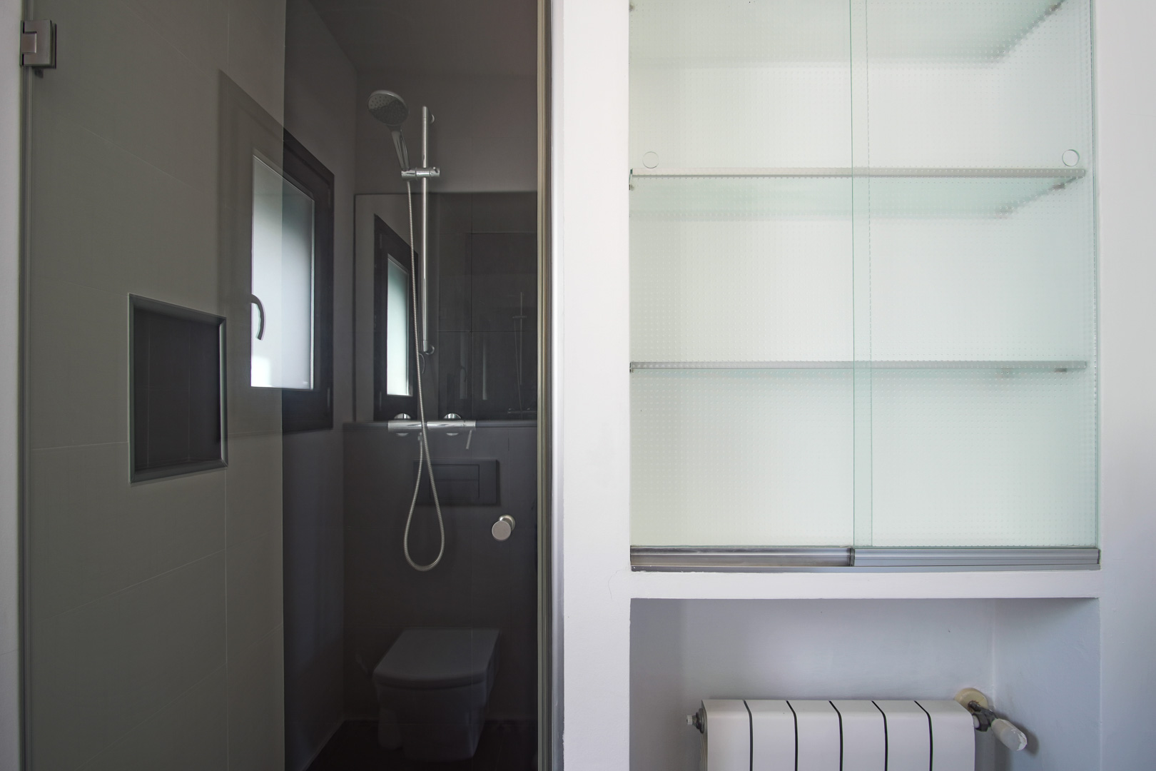 Modern bathroom featuring dark grey tiling, a walk-in shower with chrome fixtures, and a built-in storage unit with glass shelves and sliding doors.