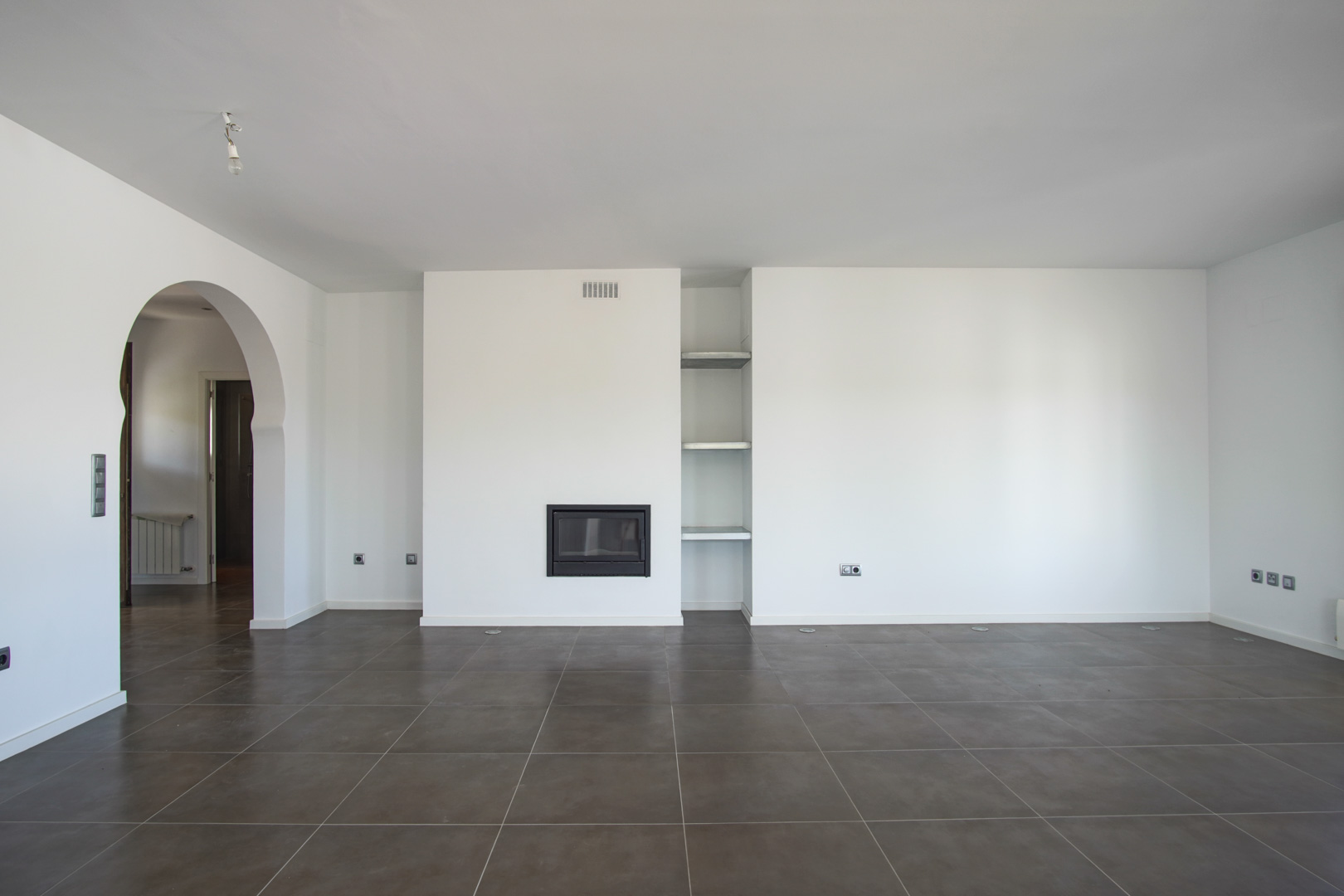 Open-plan living area featuring a built-in fireplace, recessed shelving, and dark tiled flooring. A Mediterranean archway leads to the adjacent hallway and rooms.