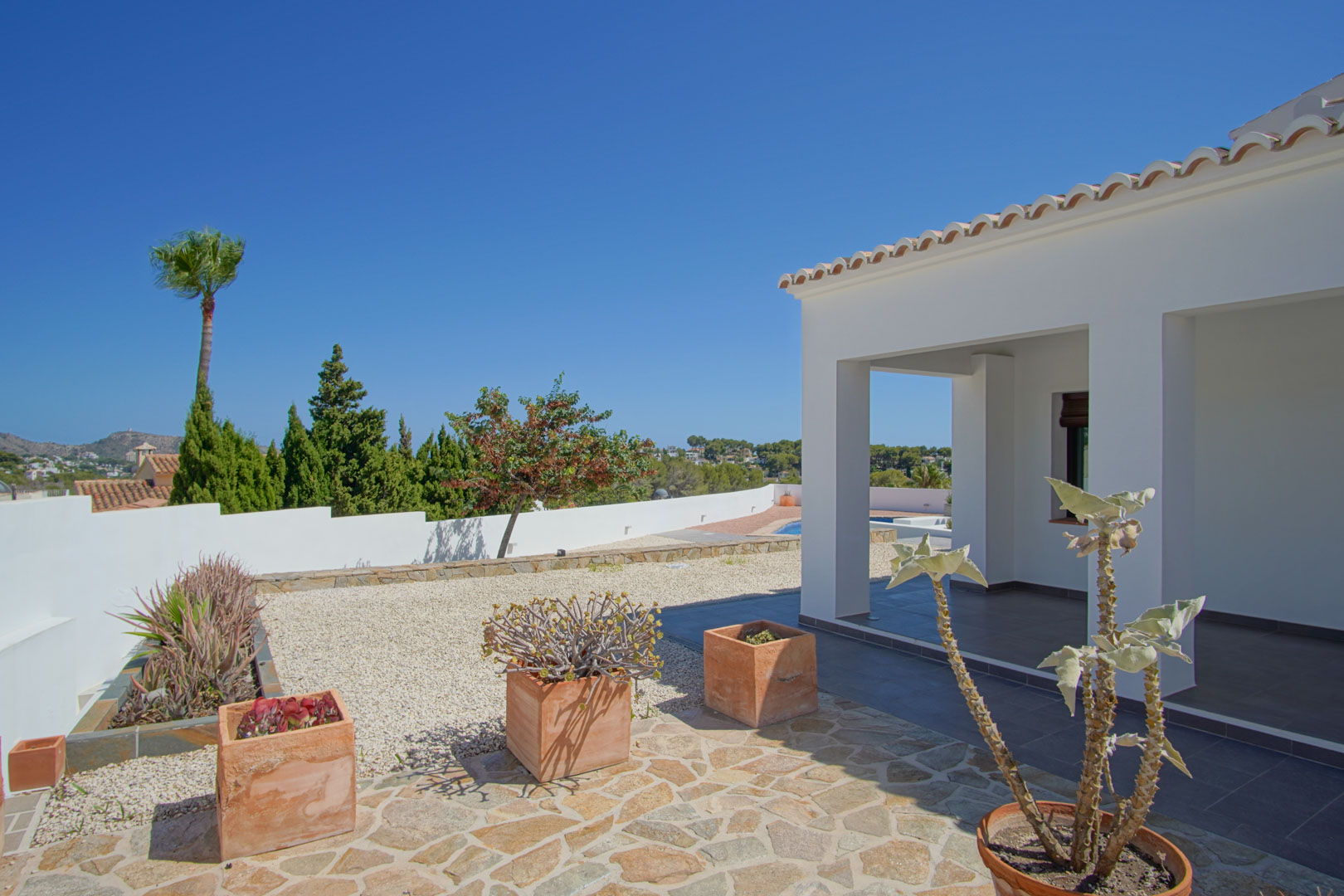 Outdoor terrace featuring a covered porch with white pillars, natural stone paving, and terracotta planters. The landscaped garden overlooks the Mediterranean coastline.