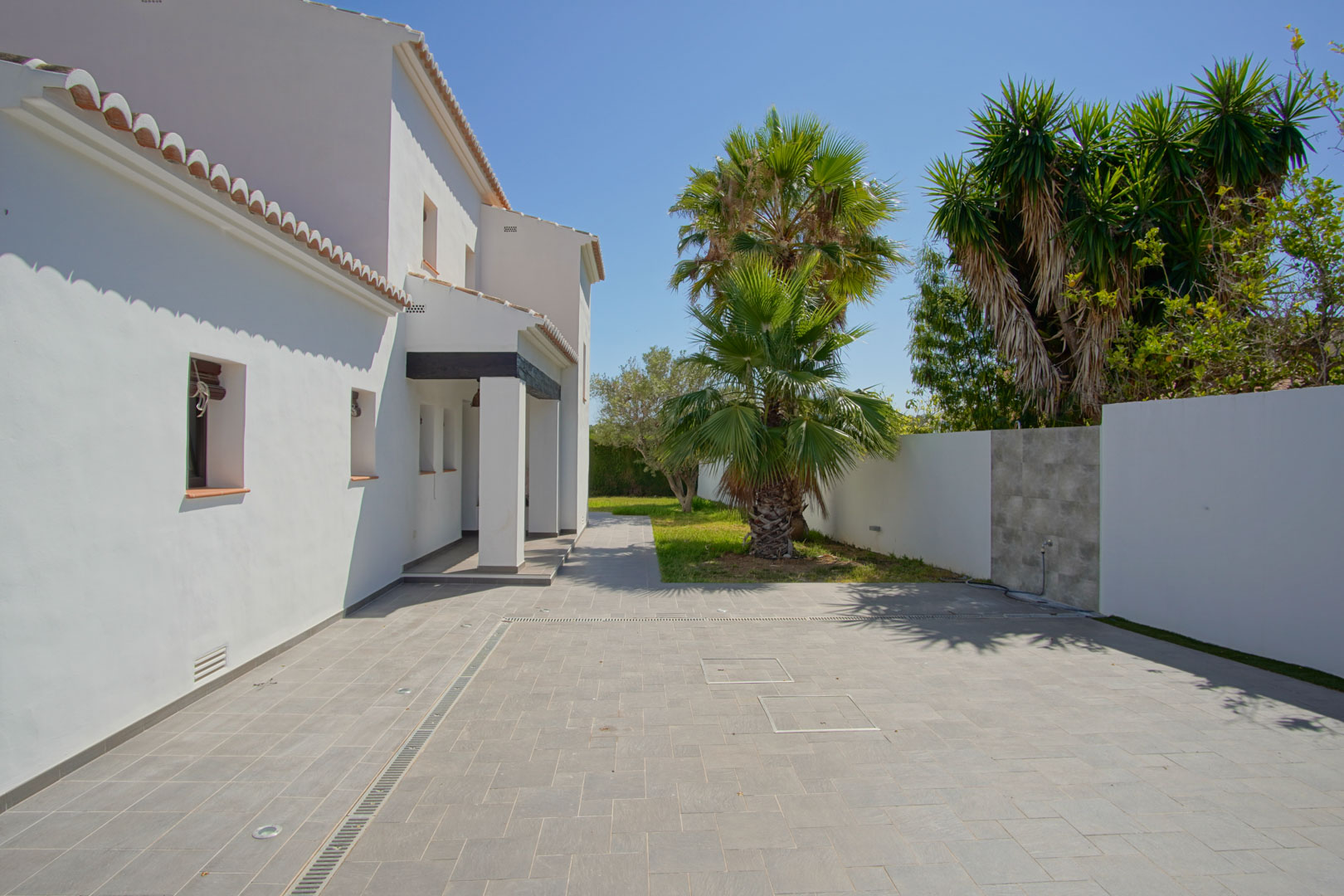 Paved entrance area featuring grey tiling, recessed floor lighting, and a Mediterranean garden with palm trees alongside the villa's white facade.
