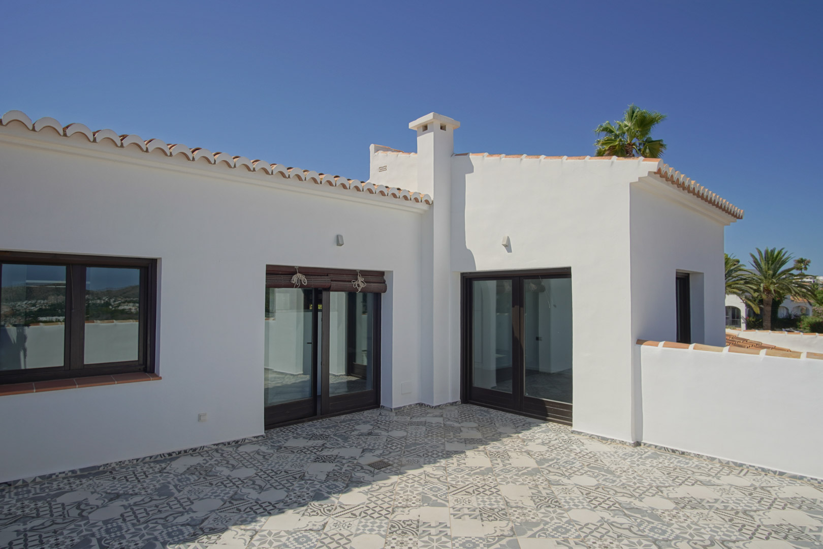 Solarium terrace featuring patterned hydraulic-style floor tiles, wood-effect aluminum joinery, and traditional Mediterranean white-washed walls.