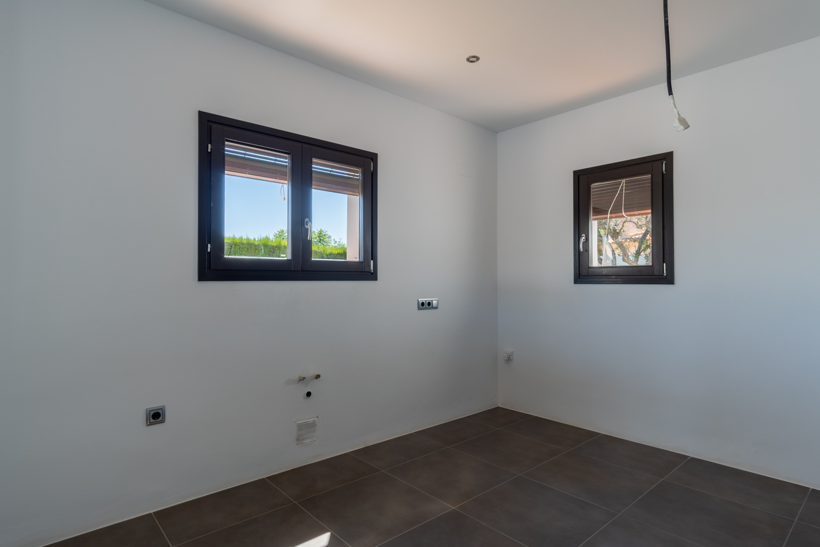 Space prepared for kitchen installation featuring dark ceramic flooring, wood-effect aluminum windows, and integrated plumbing and electrical connections.