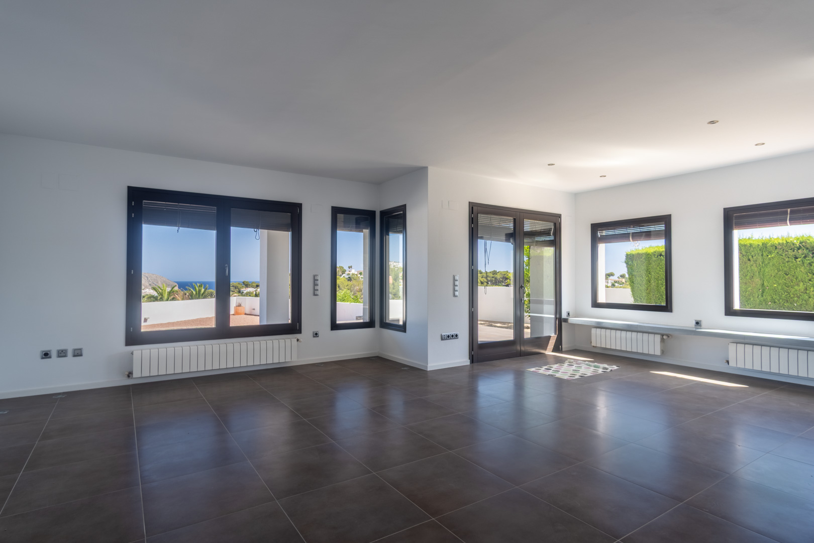 Spacious open-plan living area featuring dark tiled flooring, radiators, and wood-effect aluminum windows offering panoramic sea and coastal views.