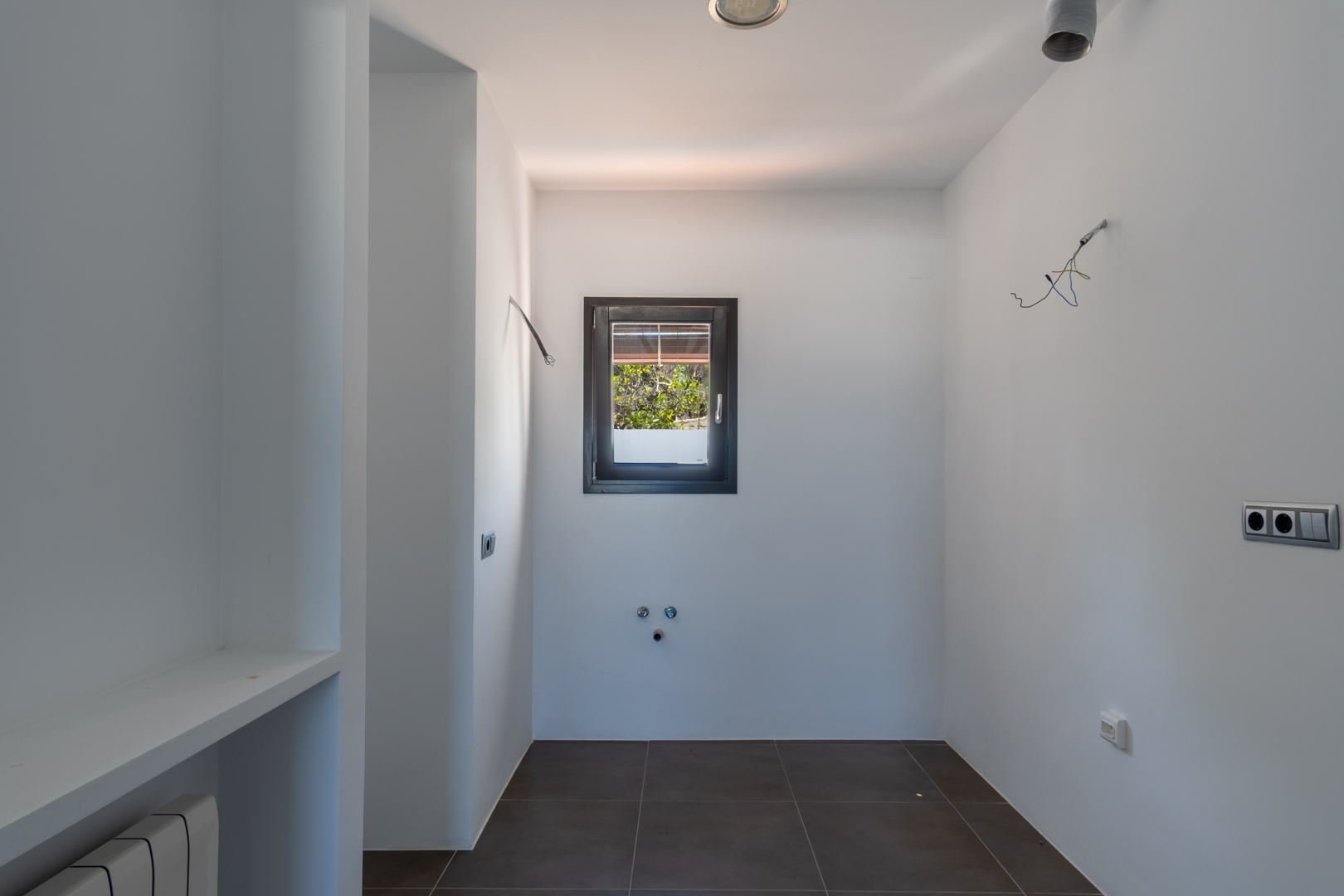 Utility room featuring plumbing and electrical pre-installations, dark ceramic floor tiles, and a dark-framed window providing natural light and garden views.
