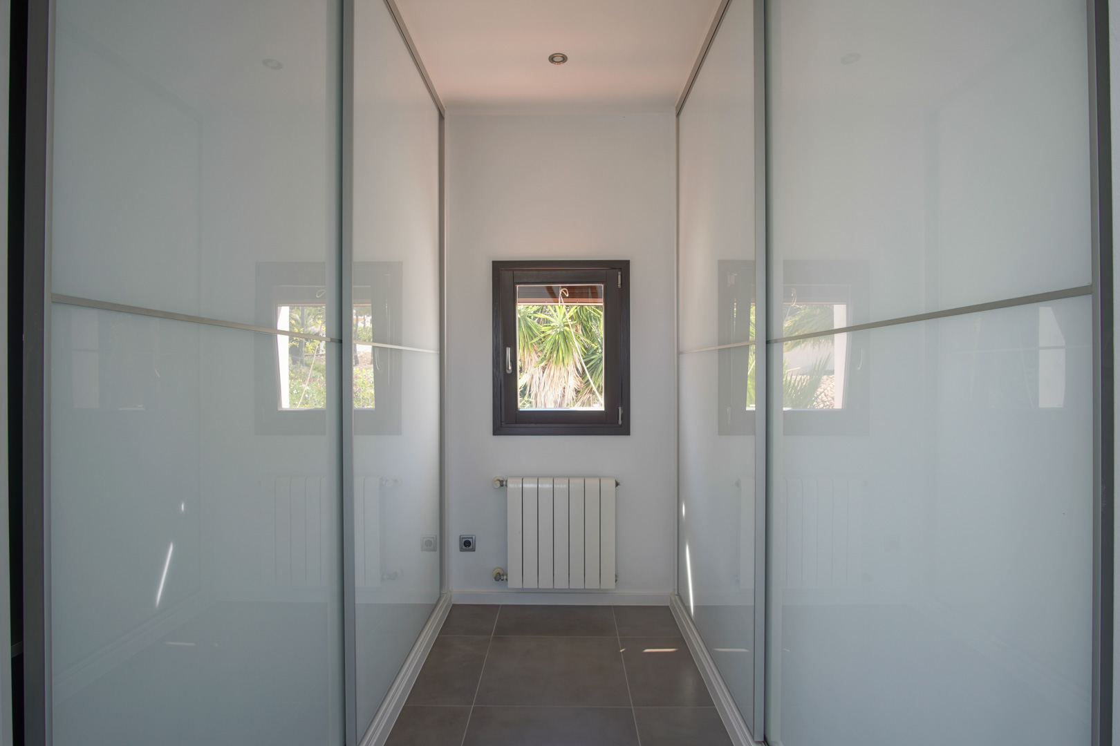 Walk-in dressing area featuring built-in wardrobes with frosted glass sliding doors, a central radiator, and a window overlooking the garden.