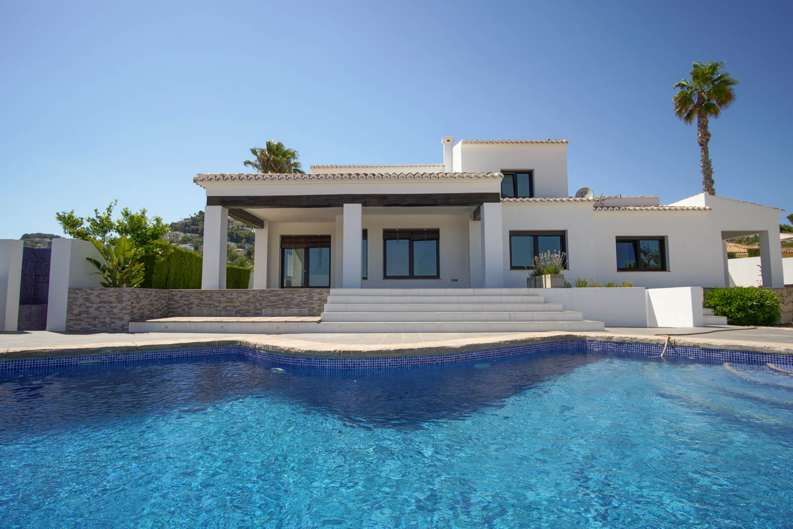 White Mediterranean villa facade with a blue tiled swimming pool. Features include a covered porch with dark wood beams and wide steps leading to the main level.