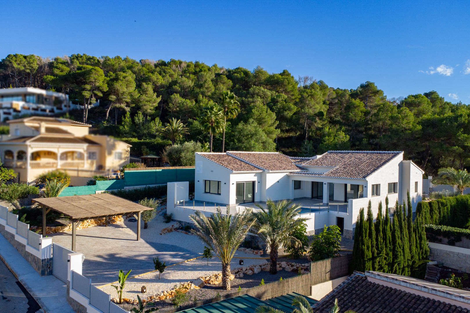 Aerial view of modern villa with brown roof tiles, white facade, and a covered carport in Jávea, surrounded by greenery.