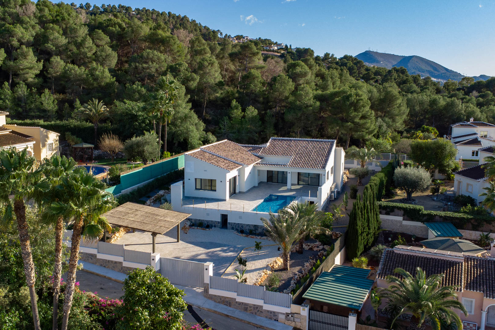 Aerial view of modern villa with pool, surrounded by lush greenery and a covered parking area in Jávea.