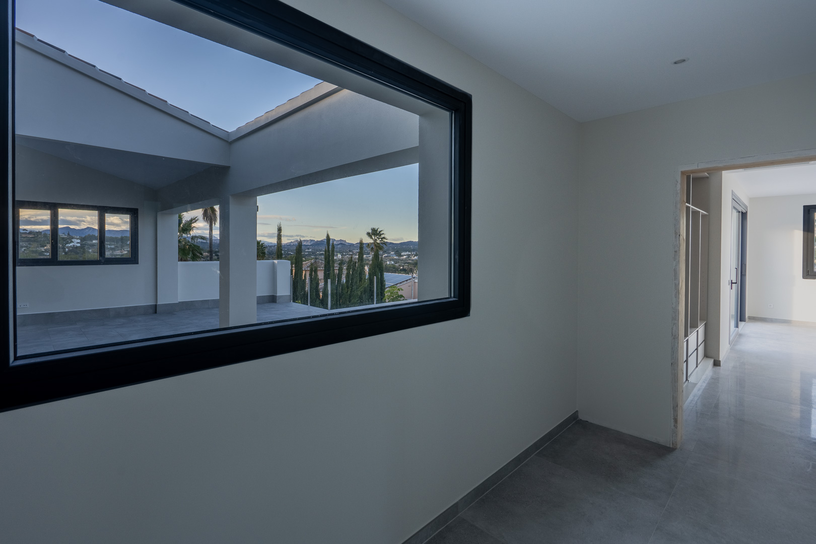 Interior view to a terrace with white railing and city view through a black-framed window.