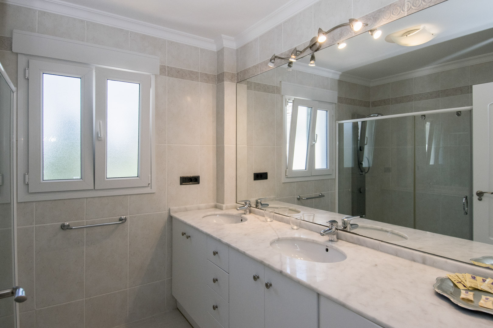 Bathroom featuring a double marble vanity, white cabinetry, and a large mirror with overhead lighting. Neutral tiled walls and a frosted tilt-and-turn window complete the space.