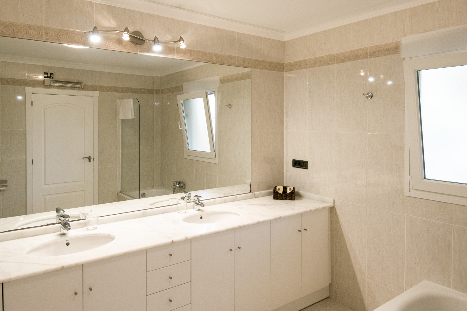 Bathroom featuring a white marble double vanity, a large wall-to-wall mirror, and integrated spotlights. The walls are finished with neutral tiles and a decorative border.