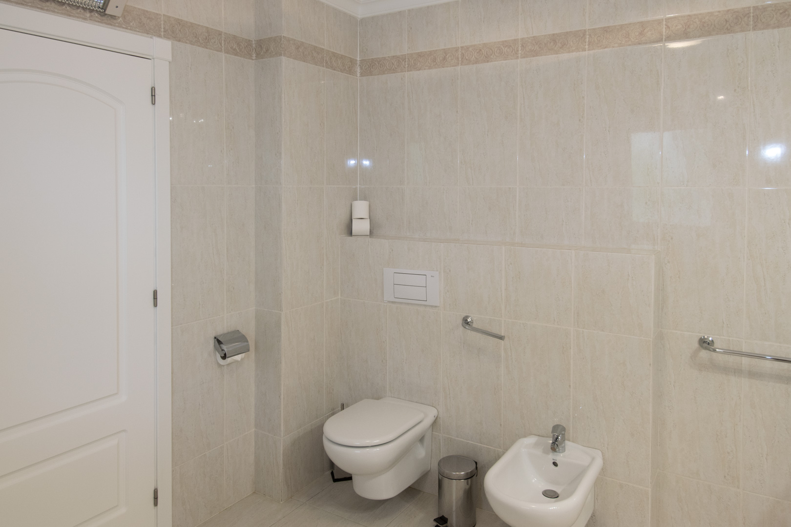Bathroom featuring cream-toned ceramic tiling with decorative borders, a wall-hung toilet, bidet, and integrated chrome safety handrails.