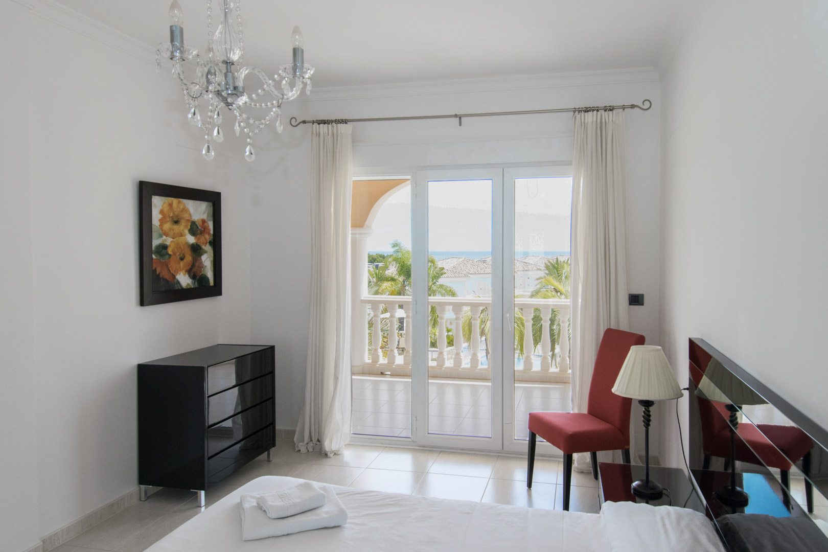 Bedroom with direct terrace access, modern black dresser, and sea views. Features include a crystal chandelier, tiled flooring, and a red accent chair.