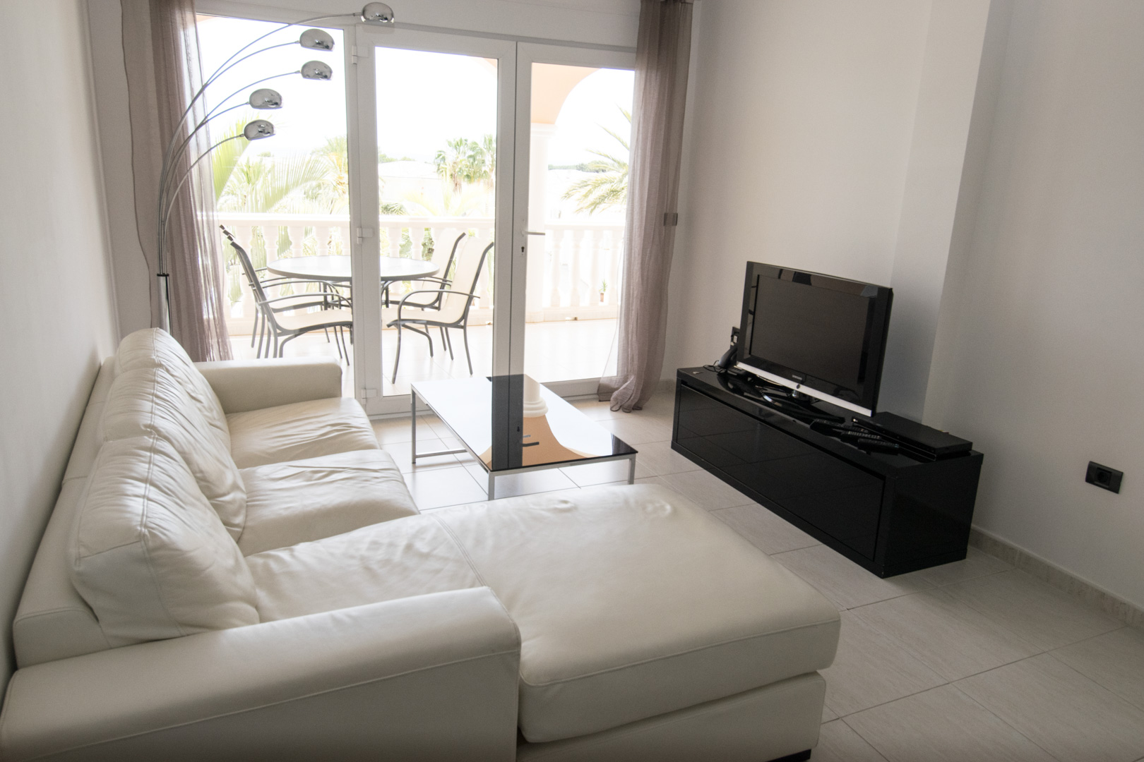 Bright living area featuring a white leather chaise longue, minimalist black media console, and floor-to-ceiling glass doors opening to a terrace.