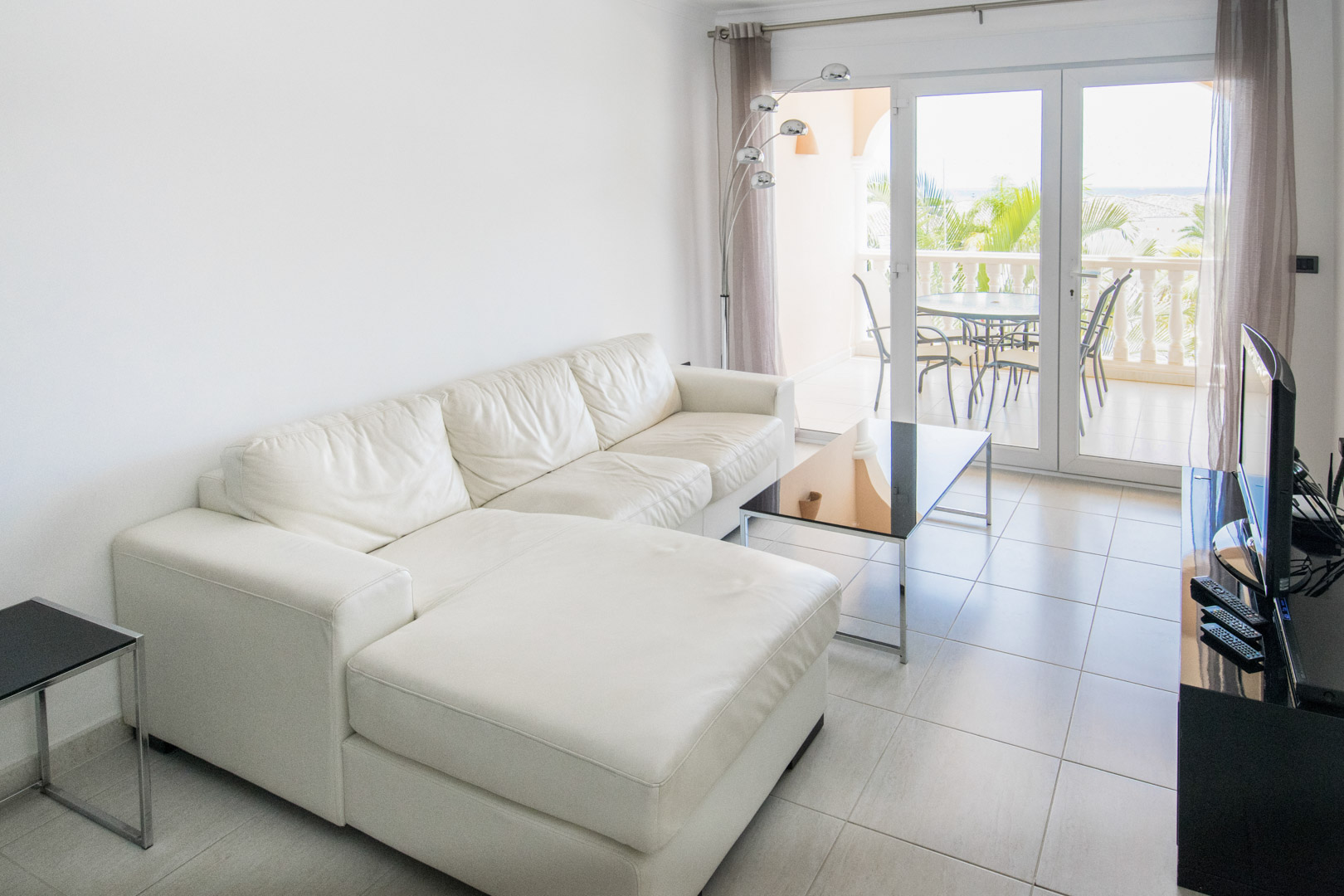 Bright living area featuring a white leather chaise longue sofa, tiled flooring, and glass doors opening onto a private terrace with sea and garden views.