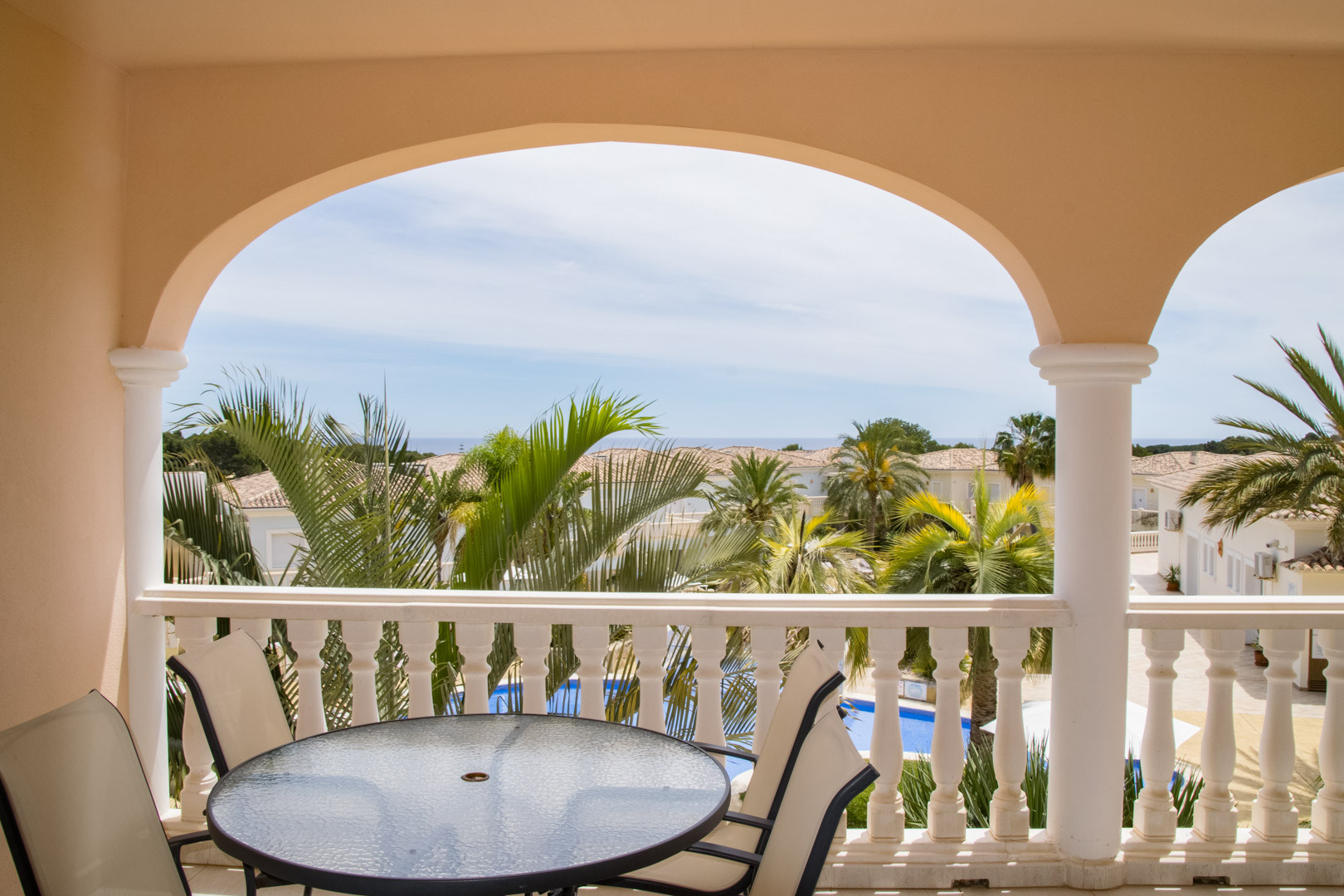Covered terrace featuring arched architecture and stone balustrades. The space includes a dining set with views over the palm-filled gardens, pool, and Mediterranean Sea.