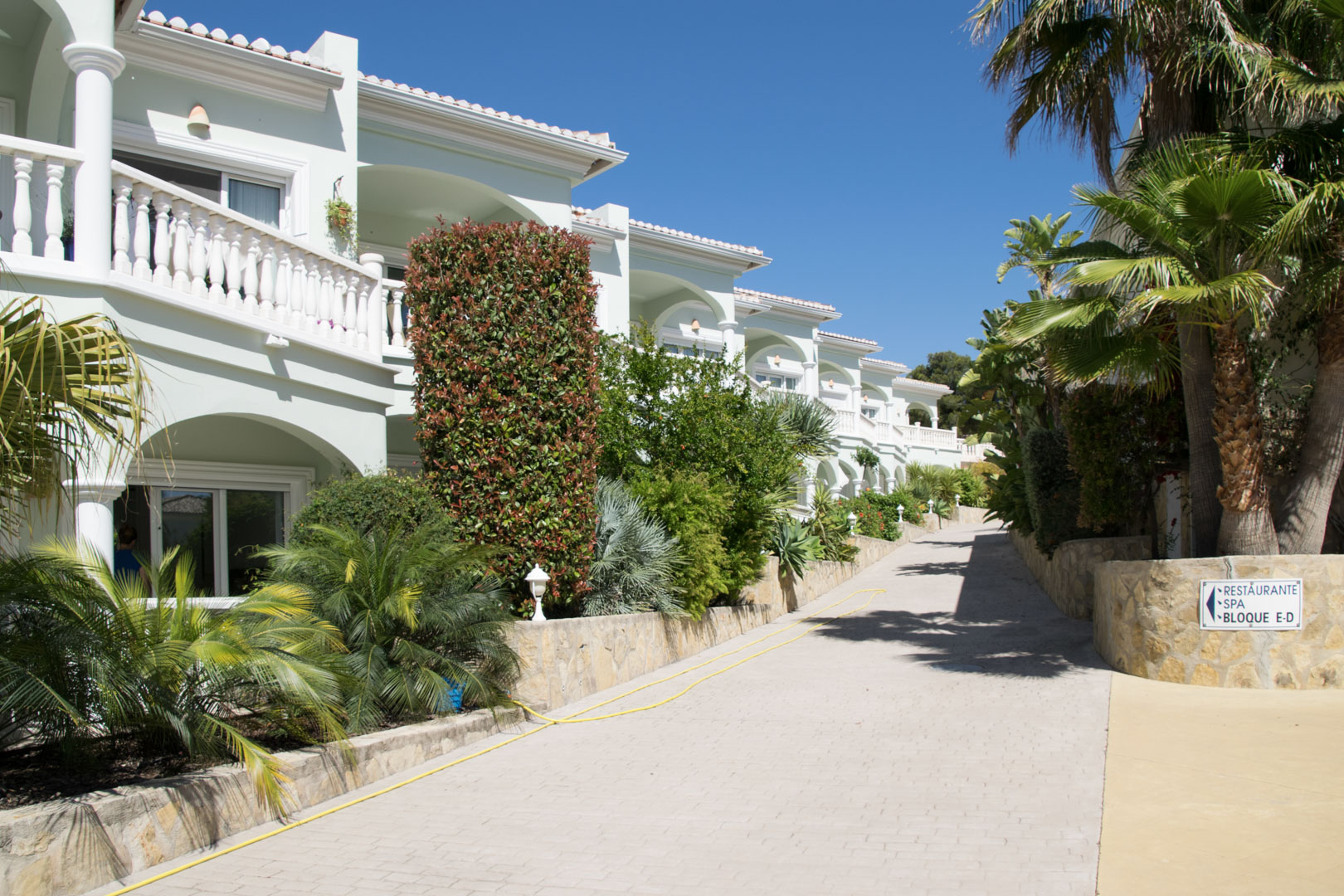 Exterior view of the Mediterranean-style complex featuring white balustrades, arched terraces, and lush landscaping along a clean, paved access path.
