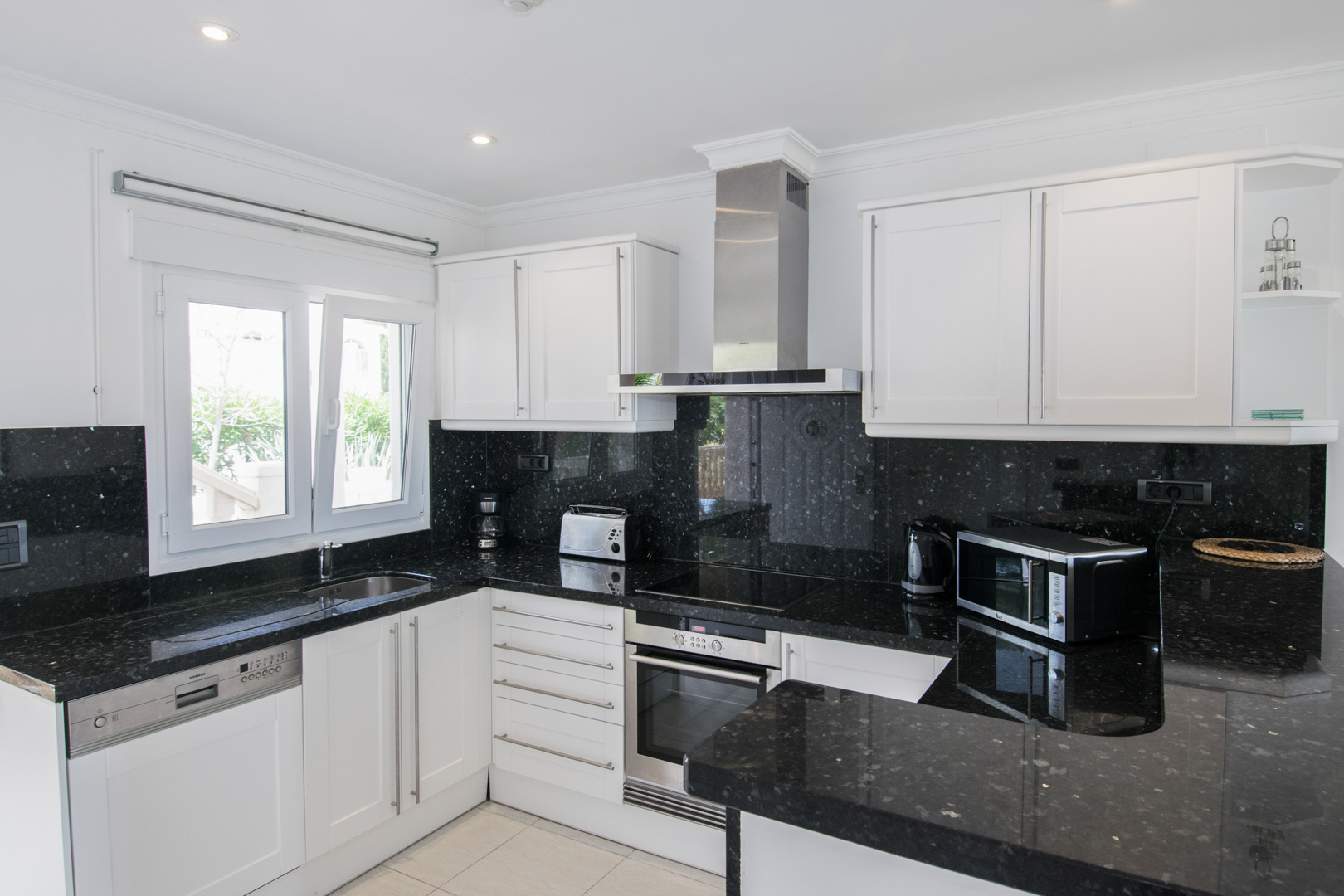 Fully equipped kitchen with black granite countertops and white cabinetry. Features integrated appliances, a stainless steel extractor, and a window providing natural light.