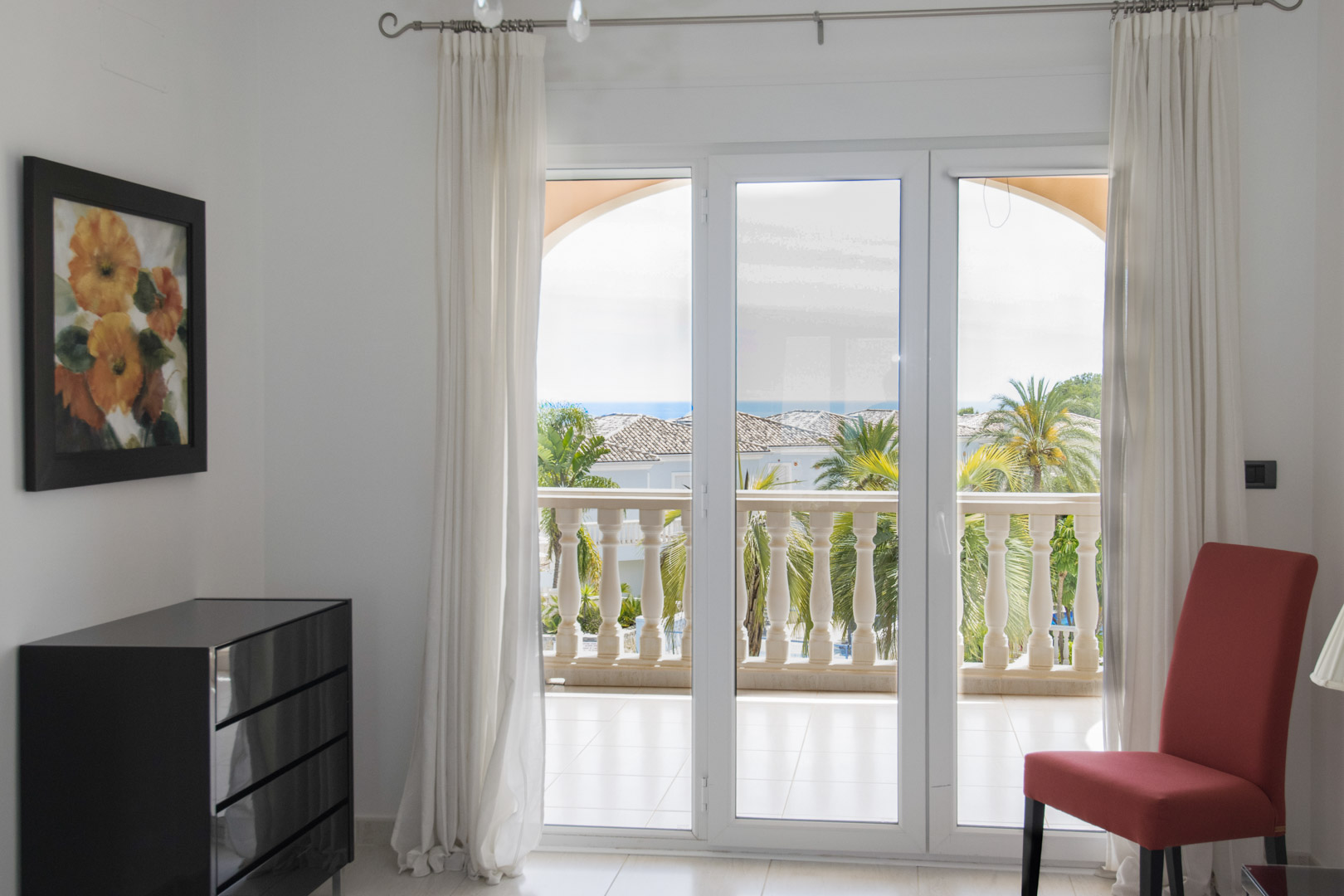 Interior view showing large glass doors opening onto a covered terrace with stone balustrades, Mediterranean gardens, and distant sea views.
