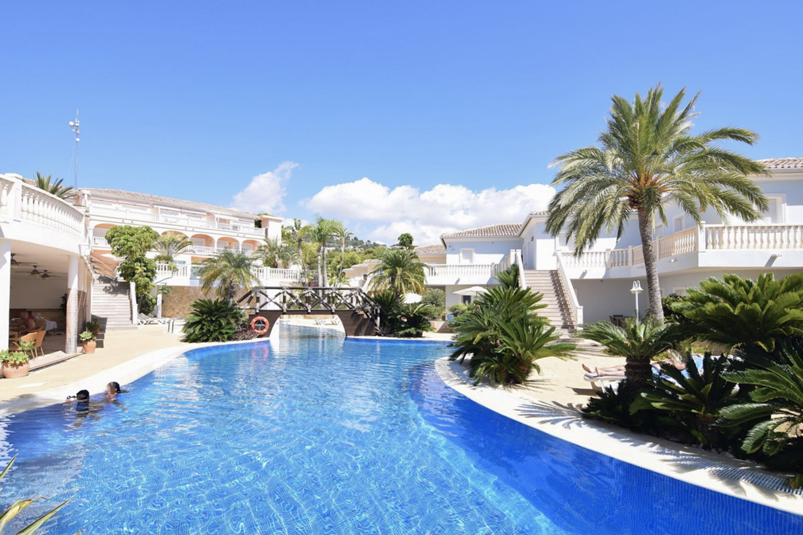 Lagoon-style communal pool featuring a wooden footbridge, surrounded by palm trees and Mediterranean architecture with white balustrade balconies.