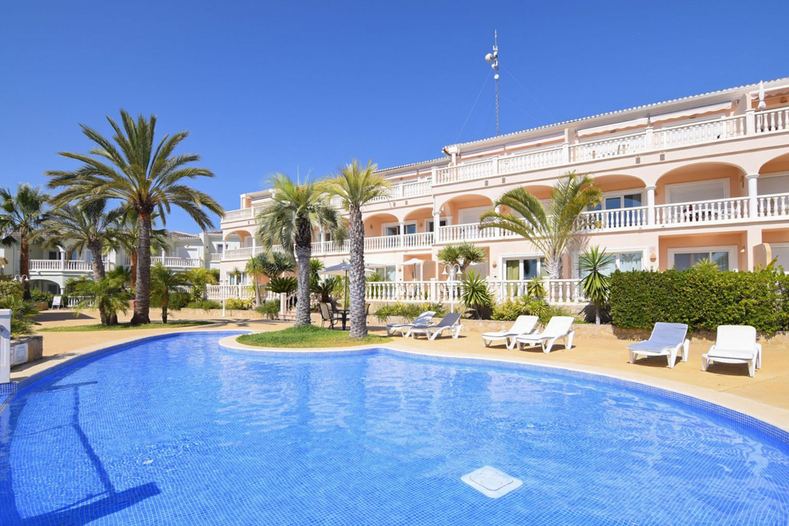 Lagoon-style communal pool featuring blue tiling, surrounded by palm trees and sun loungers against a Mediterranean building with arched terraces and white balustrades.