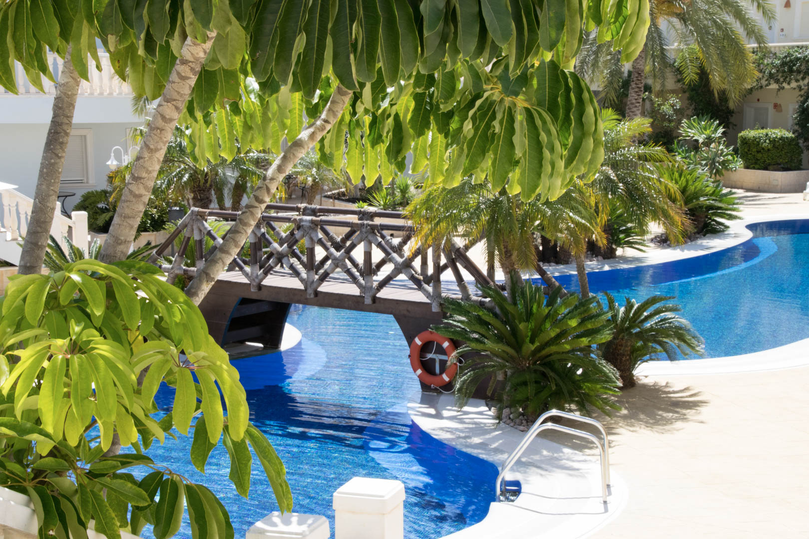 Lagoon-style swimming pool featuring a decorative wooden bridge, surrounded by lush Mediterranean gardens and tropical palm trees.