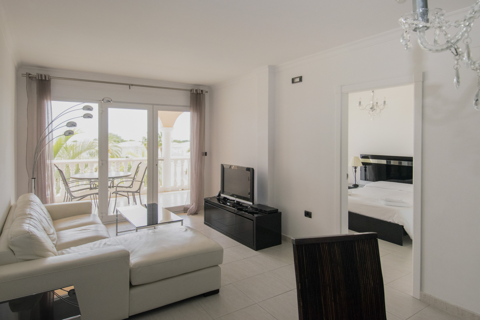 Living room featuring a white leather sofa, black minimalist furniture, and terrace access. The space includes crystal chandeliers and views into the adjacent bedroom.