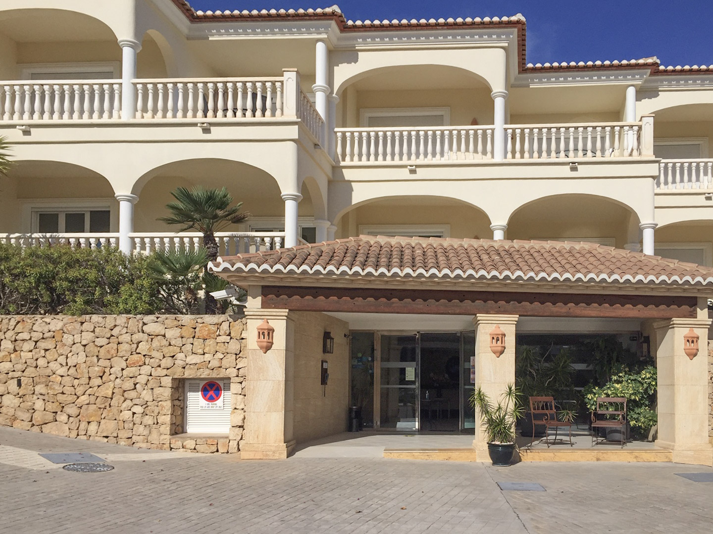 Main entrance featuring a timber-beamed porch, natural stone walls, and Mediterranean architecture with arched terraces and classic white balustrades.