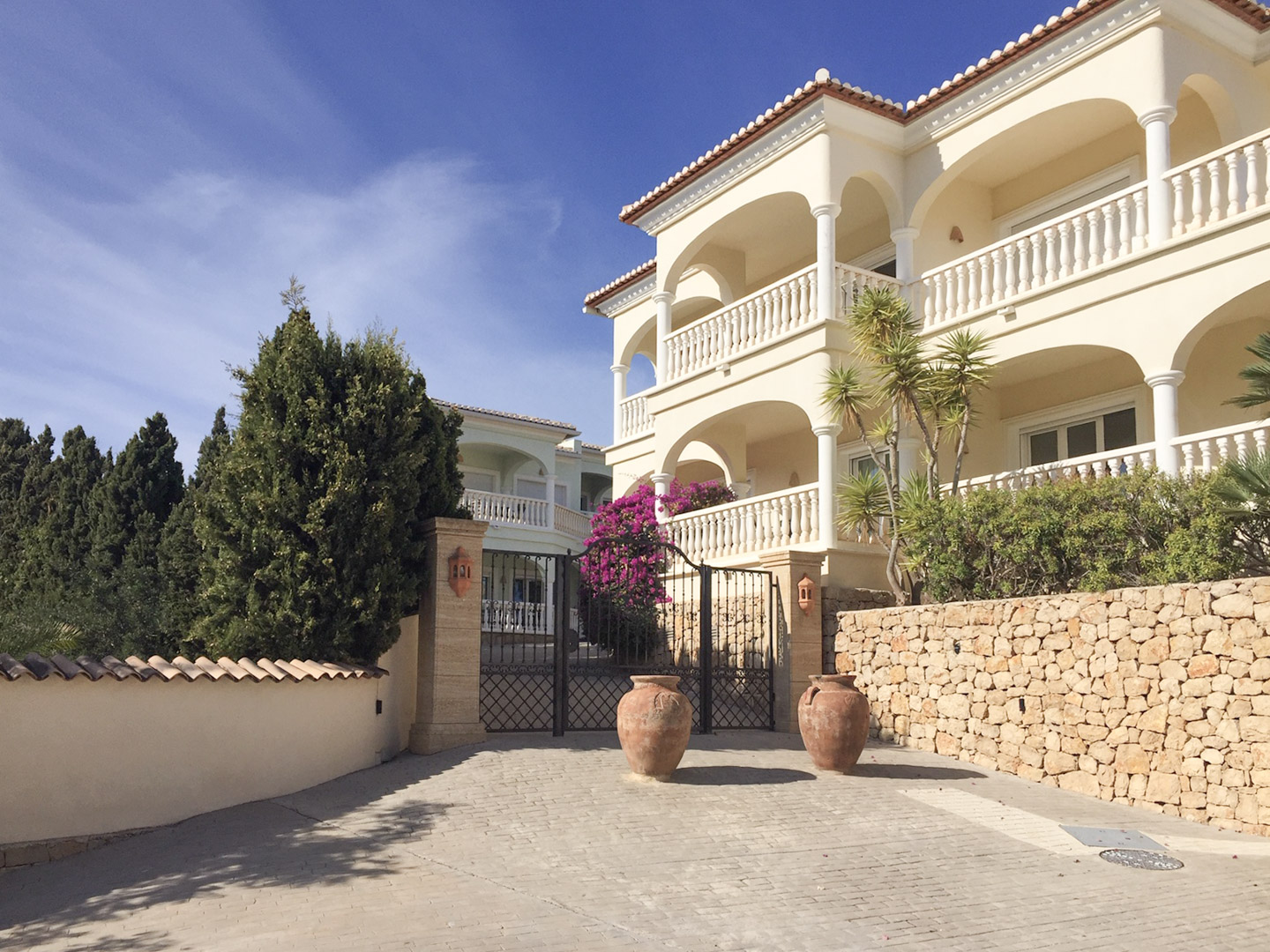 Main entrance featuring wrought iron gates, natural stone walls, and Mediterranean architecture with arched terraces and decorative terracotta amphorae.