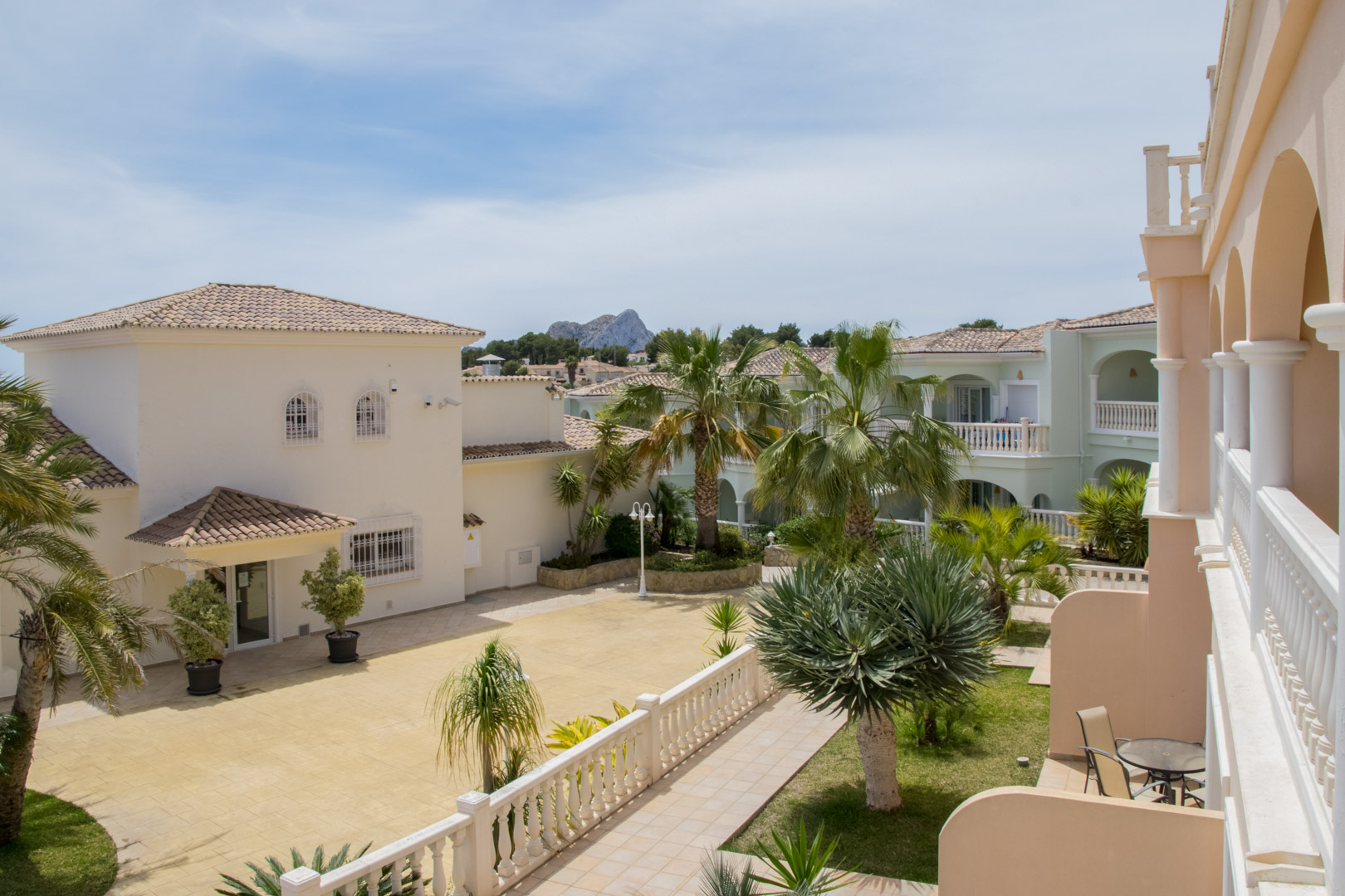 View from the complex terrace featuring Mediterranean architecture, landscaped gardens with palms, and a distant silhouette of the Peñón de Ifach.