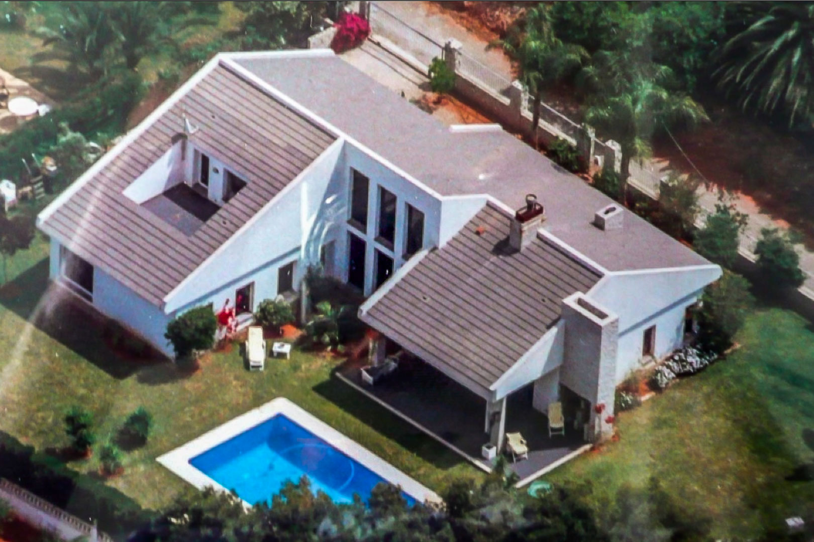 Aerial view of the white villa featuring a tiled roof, rectangular swimming pool, and manicured garden with a covered terrace and perimeter fencing.