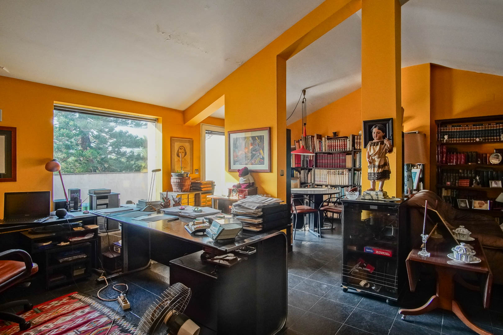 Attic study featuring sloped ceilings, mustard-colored walls, a large desk, and integrated bookshelves, illuminated by natural light from a large window.