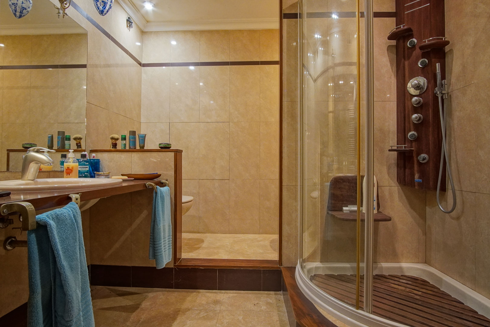 Bathroom featuring beige marble tiling, a wooden countertop, and a curved glass shower cabin with a hydromassage column and wooden slatted floor.