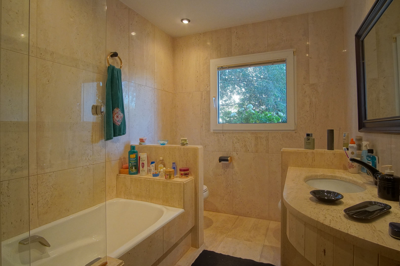 Bathroom featuring full cream marble tiling, a built-in bathtub with glass screen, and a matching stone vanity. A window provides natural light and views of the greenery.