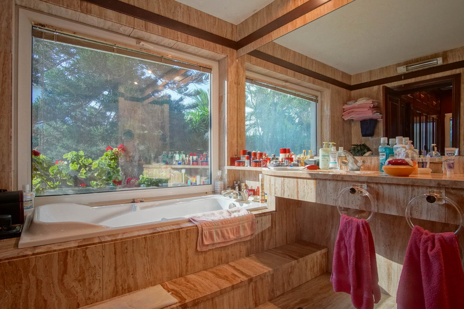 Bathroom featuring travertine marble cladding, a built-in bathtub by a large window with garden views, and an integrated vanity with a wide mirror.